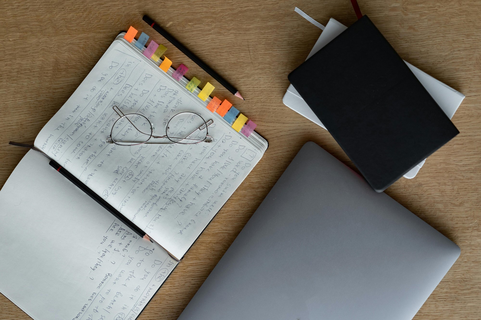 Overhead shot of a wooden desk featuring a notebook with notes, glasses, pencil, laptop, and other books.