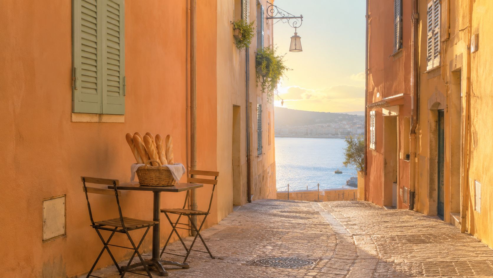 A basket of fresh baguettes on a cafe table in a narrow, sunlit cobblestone street in France.