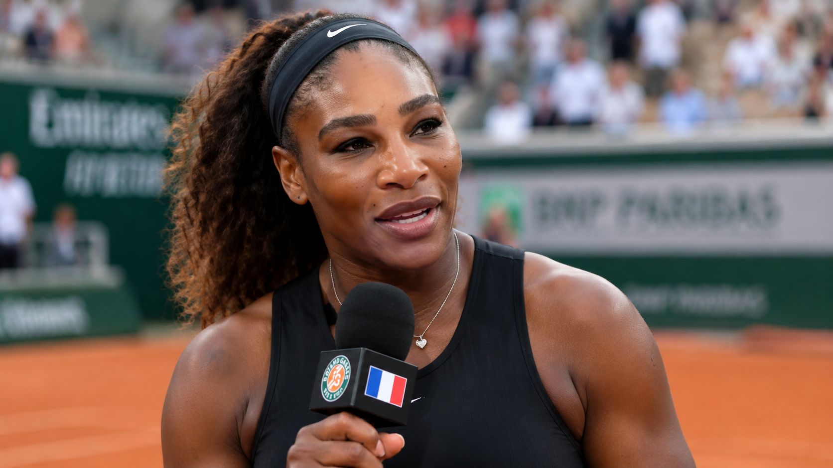 Serena Williams holding a microphone during a post-match interview on a professional clay tennis court.